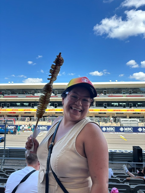 Liana at a Formula 1 race with a snack, grandstand behind her
