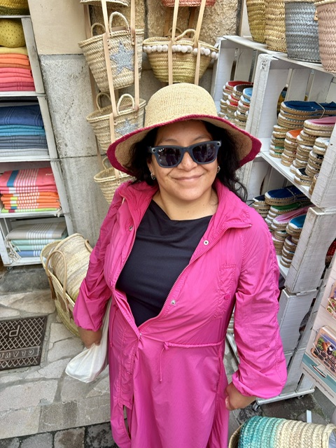 Liana in a pink jacket browsing woven baskets at a market abroad